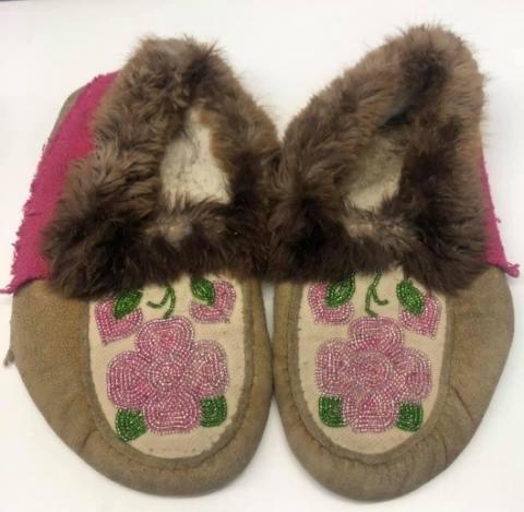 Two brown and pink hide moccasins sit side by side, with pink and green beaded flowers on the tops, and brown fur circling the opening. 