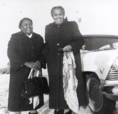 A black and white photo of two smiling women in standing in front of a car. The women's names are Lossie Lane and Ivy Beaver. The women are holding a purse and scarf. 