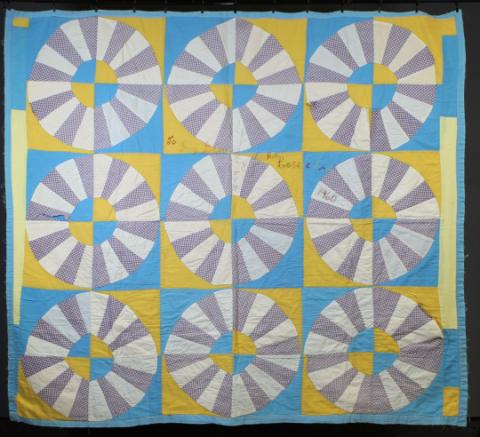 A photo of Lossie Lane's Wagon Wheel Quilt. The quilt has a pattern of white and grey wheels on top of yellow and blue squares.