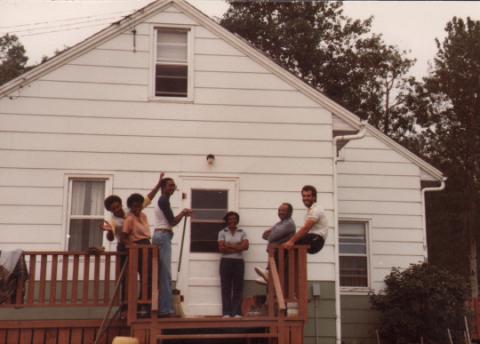 A colour photo of a happy looking family standing on the porch of a house. There are six people in the photo smiling out at the camera. 