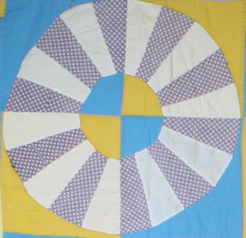 A close-up of one of the wheels from the Wagon Wheel Quilt. The wheel is made of interchanging white and grey portions. The wheels are set on squares of blue and yellow.