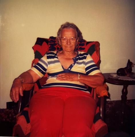 A photo of an older woman sitting in a chair, looking straight at the camera. She wears red pants, a striped shirt, and glasses. Her right hand rests on the armrest of the chair, her left rests on her stomach.