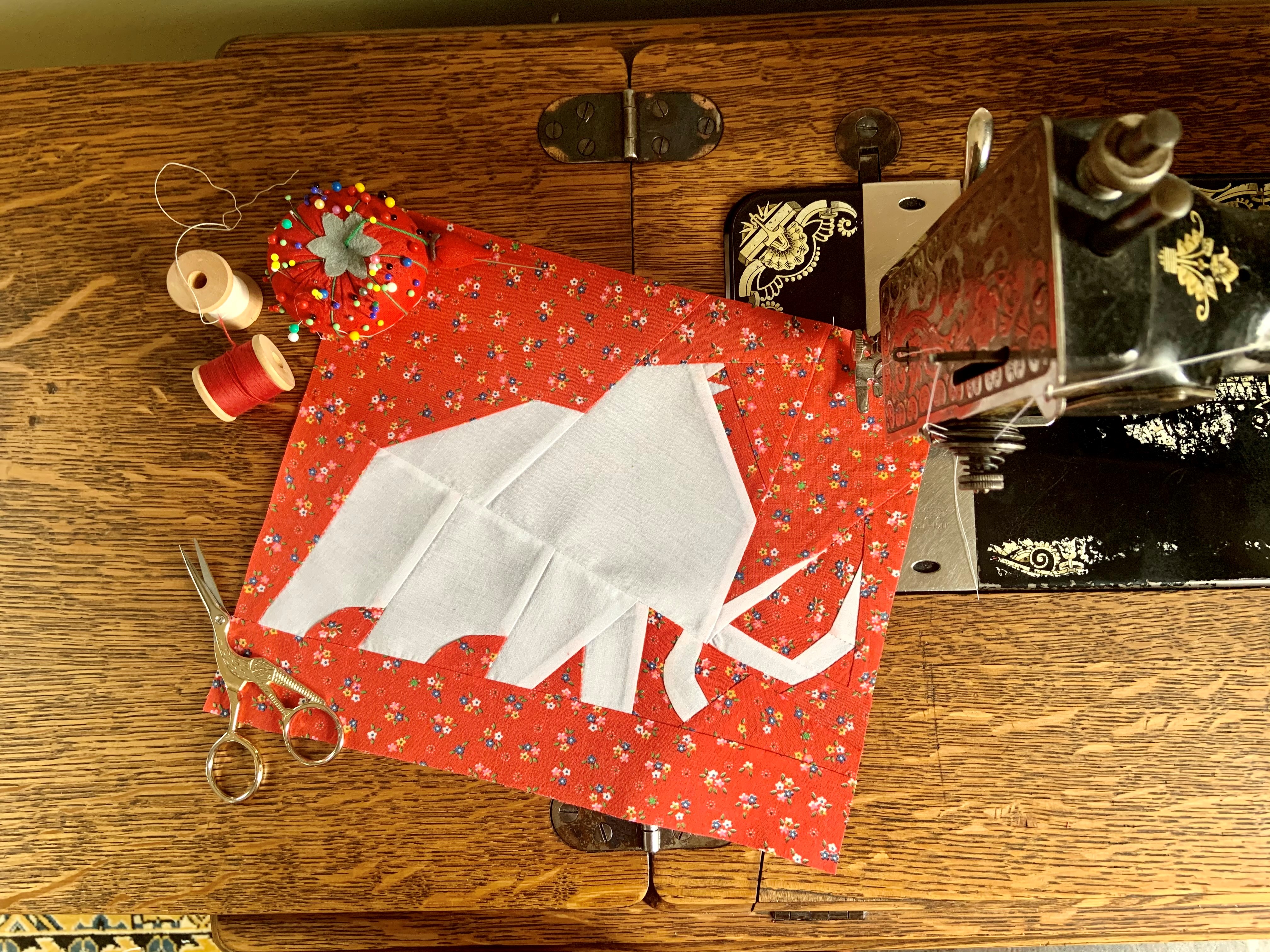 A quilted piece of fabric depicting a white mammoth lays on a wooden table next to an antique sewing machine, thread, scissors and sewing pins.