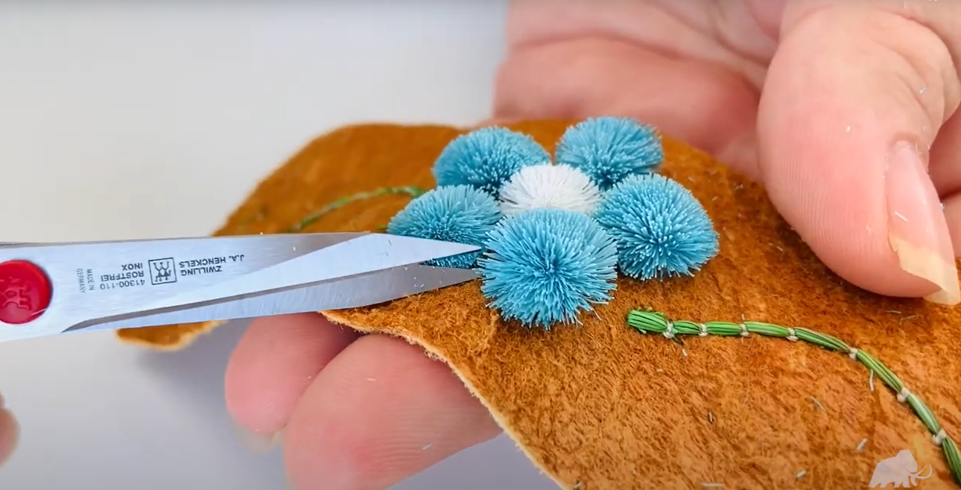 A hand holds a piece of tanned hide, with a blue caribou fur tufted flower. Silver scissors trim the tufting.