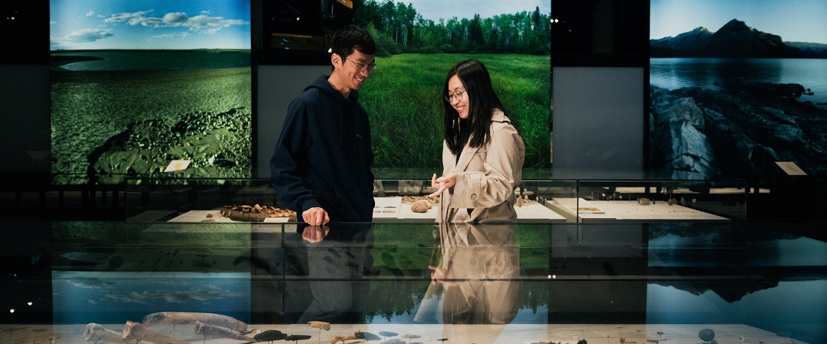 A woman and man smile as they look at artifacts inside an exhibit case. Behind them are large, illuminated photos of Alberta landscapes.