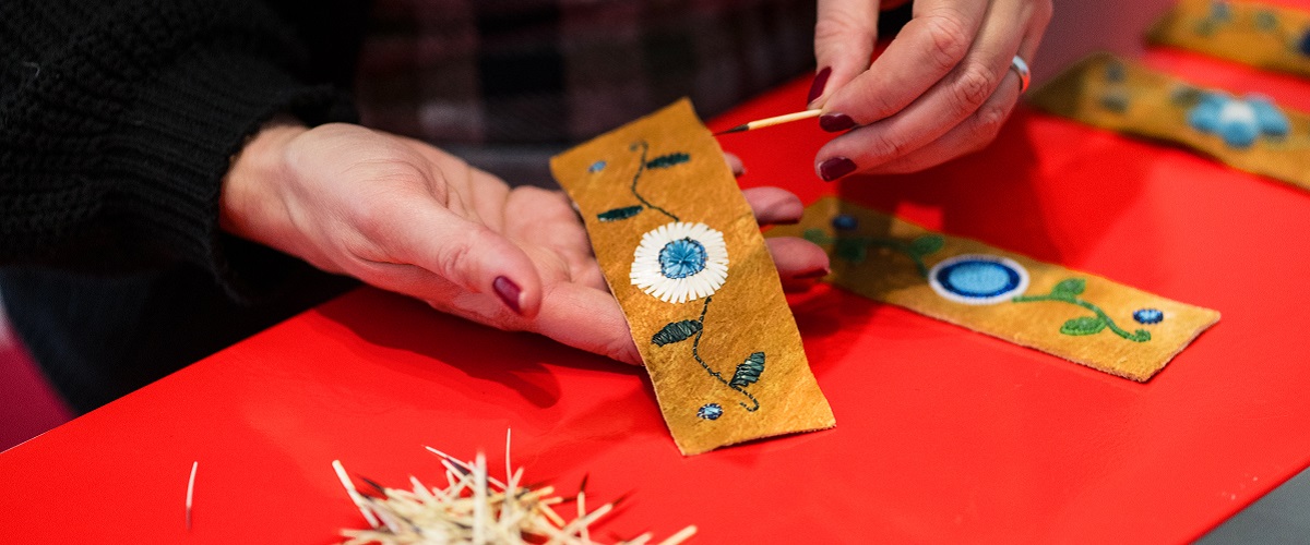 Hands hold an intricate quillwork flower design on a leather strap. Beneath the hands on a red table cloth are other quillwork designs and a small pile of quills.