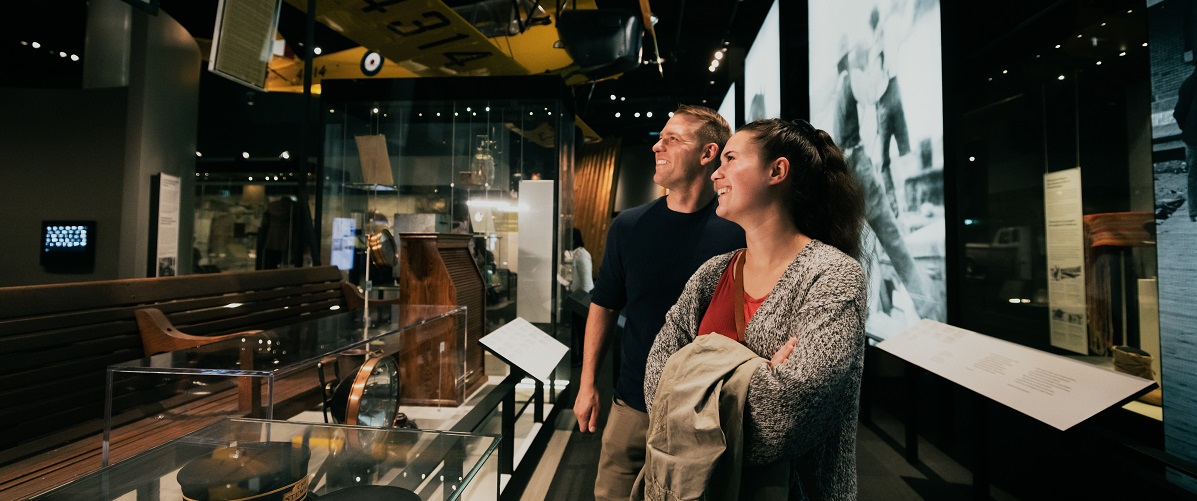 A couple smiles as they look up at a yellow airplane suspended from the ceiling in the Human History Hall