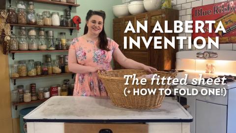 A brunette woman stands holding a woven laundry basket. The words "An Alberta Invention. The fitted sheet (and how to fold one)