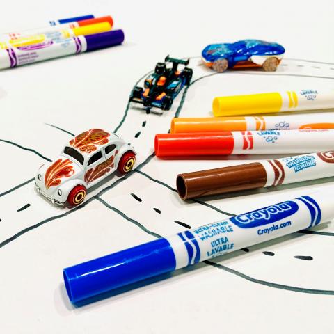 Small toy cars drive on a hand-drawn road. Felt markers in blue, brown, orange, yellow and red lay nearby.