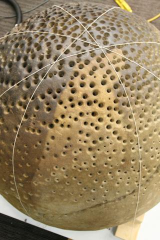 An overhead photo of the round rock with small holes. The rock is wrapped in criss-crossed string mapping out segments to be cleaned. One clean segment is a lighter brown than the rest. 