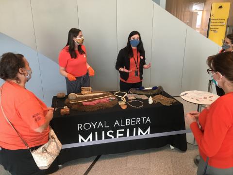 Two RAM staff educators wearing orange shirts stand behind a table, atop which sits a number of artifacts. Visitors stand facing the table