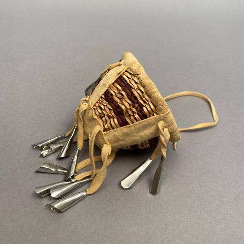 A woven basket lays on its side. The sides of the basket are woven with two alternating shades of brown. The bottom has light brown ribbons that hang, with silver metal jingles tied to the ends. 