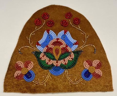 A brown piece of tanned hide featuring beaded flowers in blues, pinks and reds. The hide shape has a flat bottom and curved top.