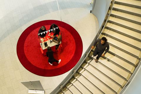 An overhead shot from the top of the main staircase in RAM. A person is walking down the staircase and you can see a group of patrons and a Museum staff member at a cart on a big round red carpet at the bottom of the stairs.