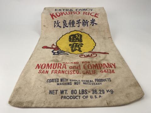 A worn rice bag with Japanese characters and the words 'Nomura and Company' lays on a white background.