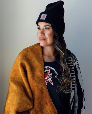 Jess Sandersson-Barry wears a hide draped over her right shoulder. She has long hair and a black toque. She is looking over her right shoulder with a peaceful smile on her face.