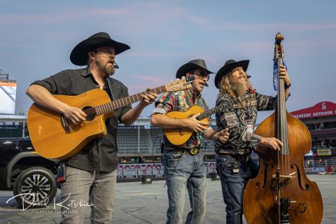 Three men in western wear and hats perform music. The man on the left has a short beard and is playing guitar, the man in the middle has sunglasses on and is also playing guitar. The man on the right has long hair and a long beard and is playing an upright bass. 