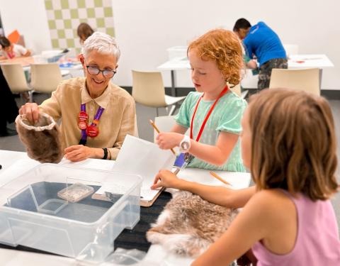 A RAM staff member sits beside two kids at a table filled with crafting supplies. The instructor is helping the kids complete summer camp projects.