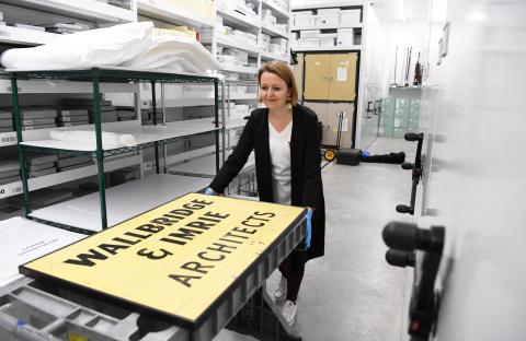 RAM Curator Jillian Richardson pushes a cart through a collections storage space. On top of the cart is a yellow sign with the words 'Wallbridge and Imrie Architects' in black text.