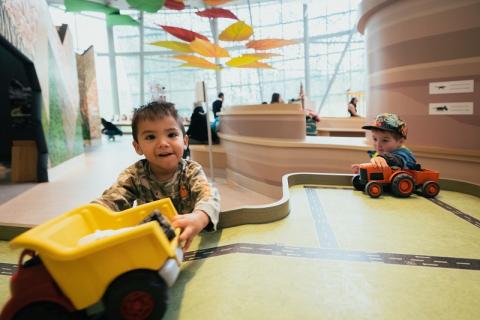 A young child plays with a large yellow toy truck in the foreground. Another young child plays with an orange toy tractor in the background. Both children are at the road table in the Children's Gallery at the Royal Alberta Museum.