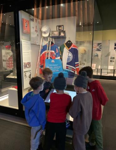 A group of five students stand around an exhibit case featuring objects related to the Edmonton Oilers hockey team.