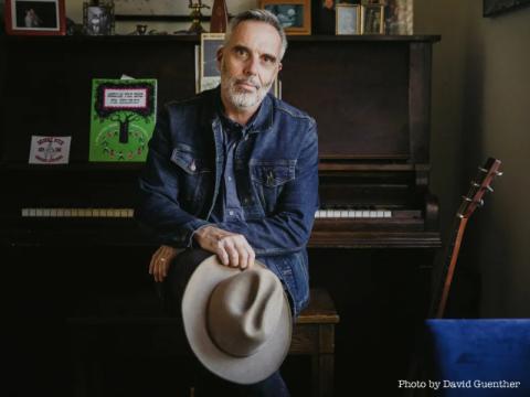 A photo of John Wort Hannam sitting in front of a piano, holding his cowboy hat.