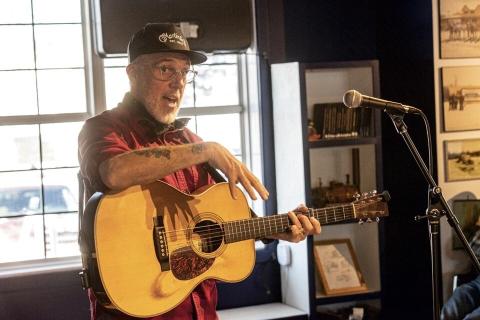 John Wort Hannam with his guitar, talking at a microphone.