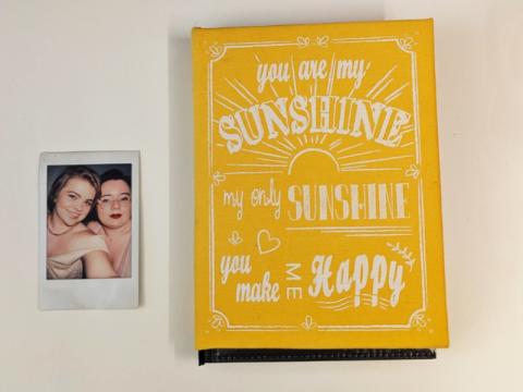 A small polaroid photo of Harper-Hugo and Will from their wedding. Beside the photo is a yellow scrapbook. The book is closed, but we know that there are mementos from their relationship inside.