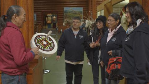 A smiling family of four adults watches an elder play a drum.