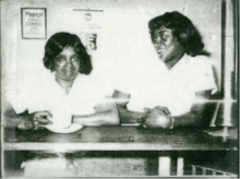 A black and white photograph of two women sitting at a table, one looking at the camera and the other looking at the other woman.