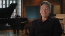 K.D. Lang looks out at the camera with a smile on her face. There is a grand piano to her side on the left side of the image.