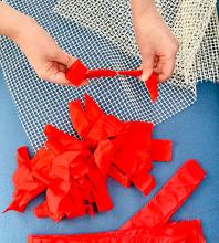 Hands hold bright red scraps of cloth. The cloth is being weaved to make a rug.