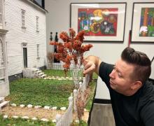 Artist/Miniaturist Lance Cardinal is crouched putting the finishing touches on the front yard of a miniature house.