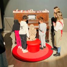 A group of children are gathered around an old wooden piano.