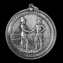 A silver medal commemorating the 1876 Treaty 6 signing. The medal depicts a Canadian treaty commissioner shaking hands with an Indigenous chief, both in formal dress, with a tomahawk between their feet and the sun and teepees in background.