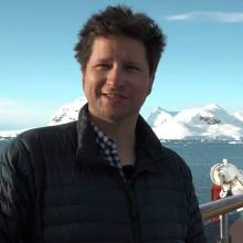 Portrait of Josh McInnes. A white man wearing a jacket is smiling out at the camera. Snowy mountains can be seen behind him in the distance.