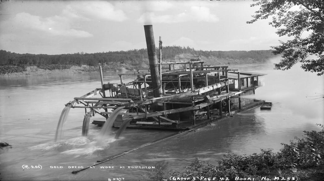 Historic black and white photo of a steam-poered mining dredge making it's way down a river.