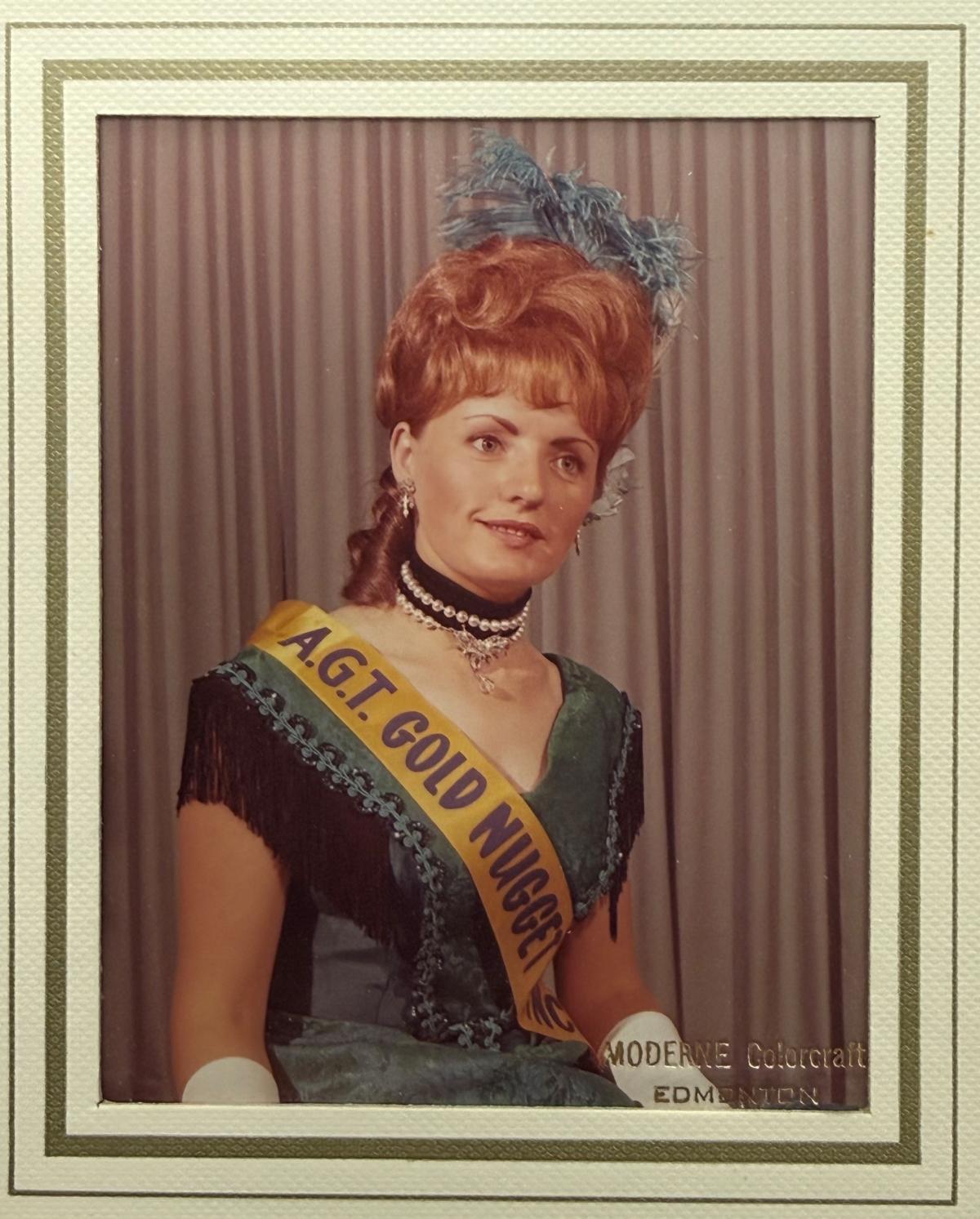 Portrait of Simone Bessette in her award winning dress. Simone smiles at the camera, she is wearing a dark green dress with black fringe accents. She is also wearing long white gloves, a green feather head piece, and a gold sash that says "A.G.T Gold Nugget Princess"