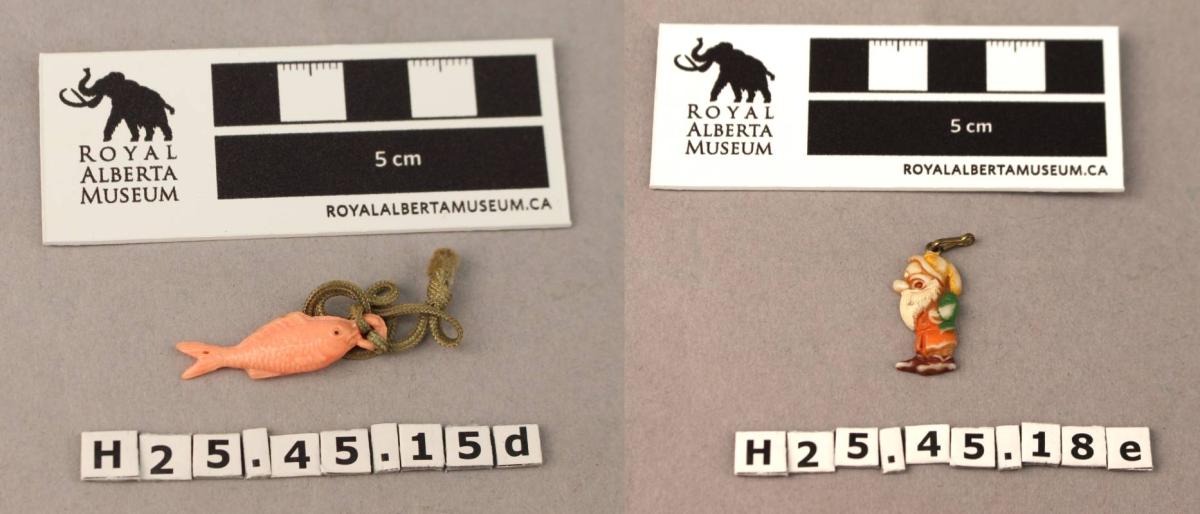 Two museum collection photos of small charms each photo has a scale reference ruler and the item number on it. The charm on the left is a small pink fish tied to a green cord. The charm on the right is a small painted, Disney-style dwarf.