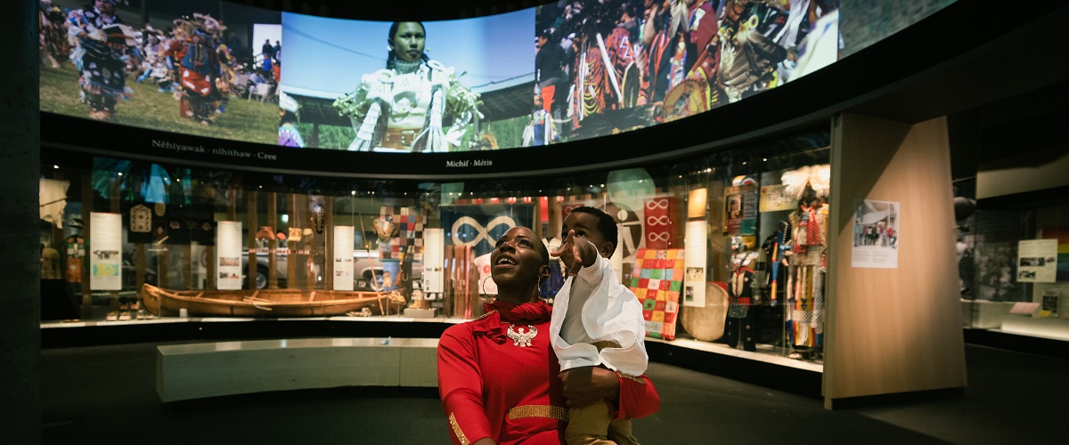 A woman and her young child sit in the circular What Makes Us Strong gallery, looking up in awe at the screens above.