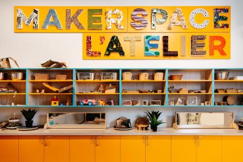 Orange shelves above an orange counter. The shelves are filled with various crafting supplies and toys. There is a colourful sign above the shelves that says Makerspace, L'Atelier